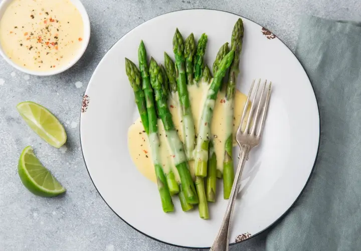 Asparagi freschi conditi con una cremosa salsa olandese, accompagnati da spicchi di lime e una forchetta d'argento.