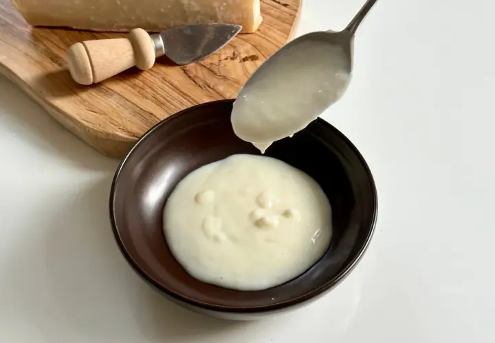 Un cucchiaio versa una densa crema di parmigiano in una ciotola. Sul tagliere, un pezzo di formaggio e un coltellino.