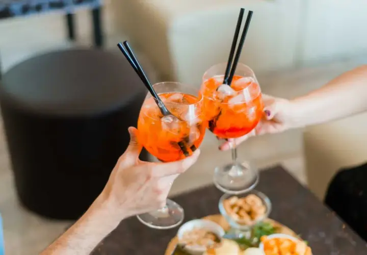 Due mani brindano con due spritz pieni di ghiaccio e cannucce nere. Sul tavolo, stuzzichini invitanti.