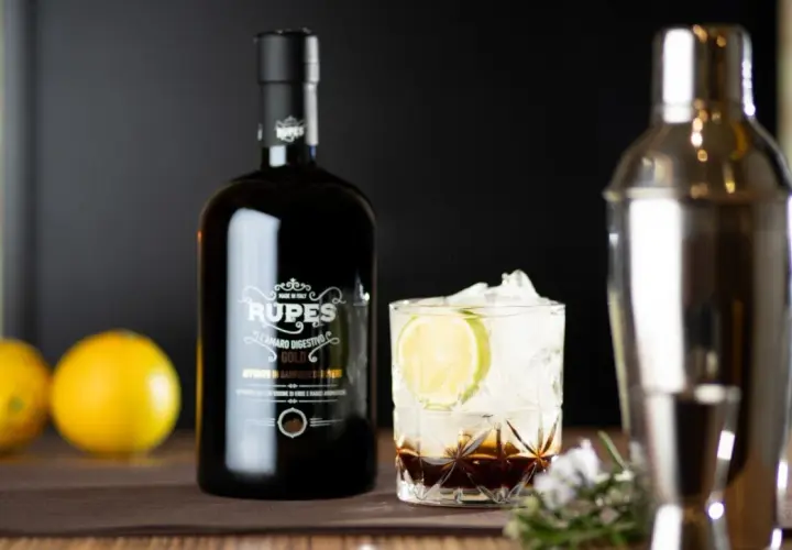 Bottiglia di amaro Rupes, un classico tra gli amari calabresi, accanto a un bicchiere con ghiaccio, lime e un cocktail.