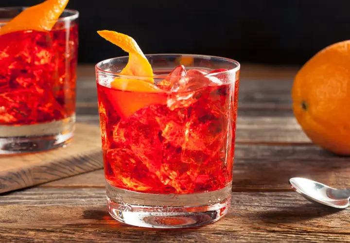 Un bicchiere di rosso vibrante, ghiaccio e scorza d'arancia. La ricetta americano è pronta per essere gustata, con un'arancia sullo sfondo.