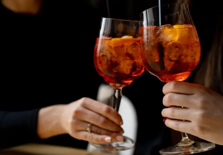 Due mani brindano con due spritz, un superalcolico rinfrescante con ghiaccio e arancia, su sfondo scuro.
