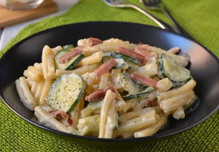 Piatto di pasta cremosa con zucchine a rondelle e cubetti di speck croccante, guarnito con pepe nero.