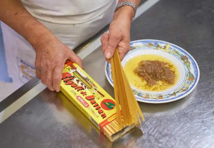 Mani che preparano i bigoli in salsa, con la scatola di pasta e un piatto di salsa di acciughe e cipolle.
