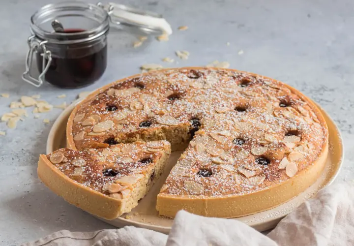 Torta con crema frangipane, mandorle a lamelle e ciliegie, spolverata di zucchero a velo. Accanto, un vasetto di confettura.