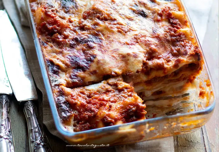 Lasagne alla bolognese appena sfornate, dorate e filanti, in una teglia di vetro. Accanto, due coltelli d'argento.