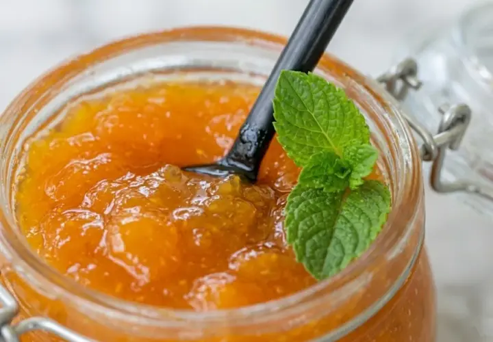 Vasetto di vetro pieno di marmellata di albicocche fatta in casa, con un cucchiaio nero e una fogliolina di menta fresca.