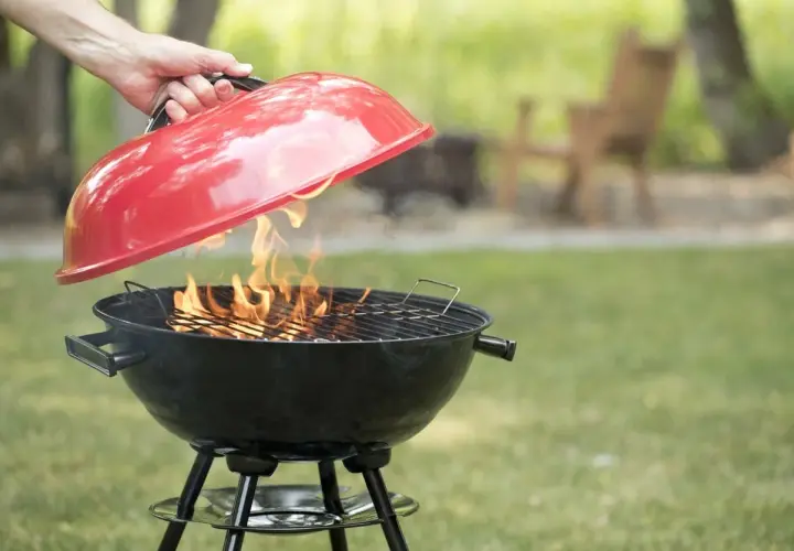 Mano solleva il coperchio rosso di un barbecue a carbonella con fiamme vive. Perfetto per chi vuole sapere se posso usare legna nel barbecue a carbonella.