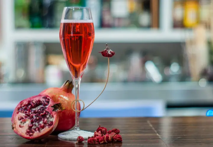 Un bicchiere di tintoretto cocktail rosso rubino, accompagnato da melograni freschi e secchi, su un tavolo di legno scuro.