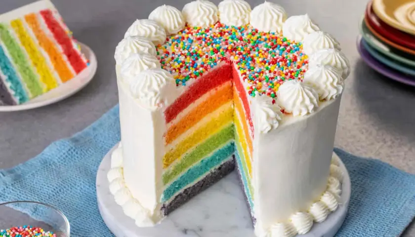 Torta arcobaleno con strati colorati, glassa bianca e zuccherini. Una fetta mostra i colori vivaci.