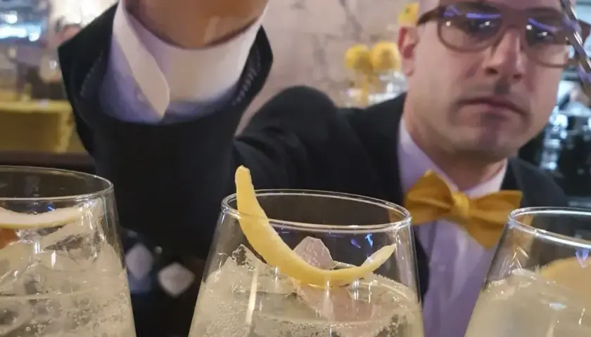 Un barista con papillon giallo prepara un bergamotto spritz, guarnito con scorza di limone.