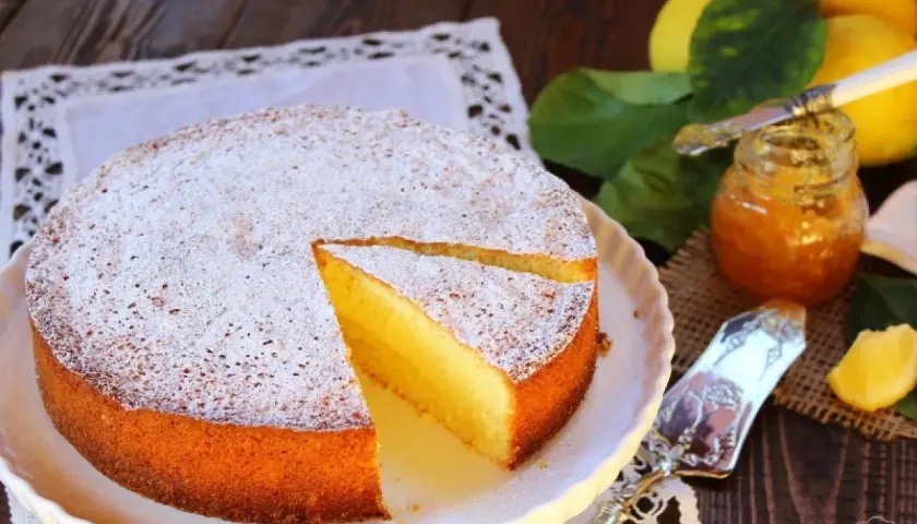 Una soffice torta al limone, spolverata di zucchero a velo, accanto a limoni freschi e marmellata.
