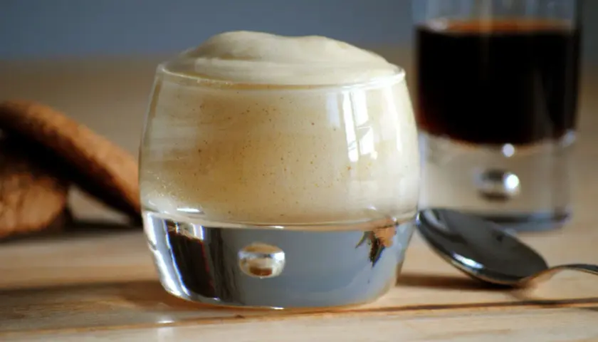 Delizioso zabaione ricetta in un bicchiere, con biscotti e caffè sullo sfondo.