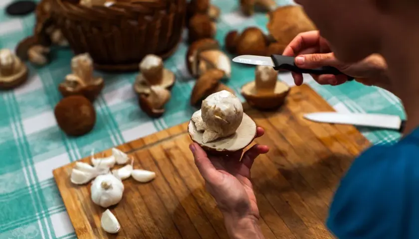 Mani che puliscono funghi freschi su un tagliere di legno, con aglio e un cesto di funghi sullo sfondo. Impara come pulire i funghi per gustarli al meglio.