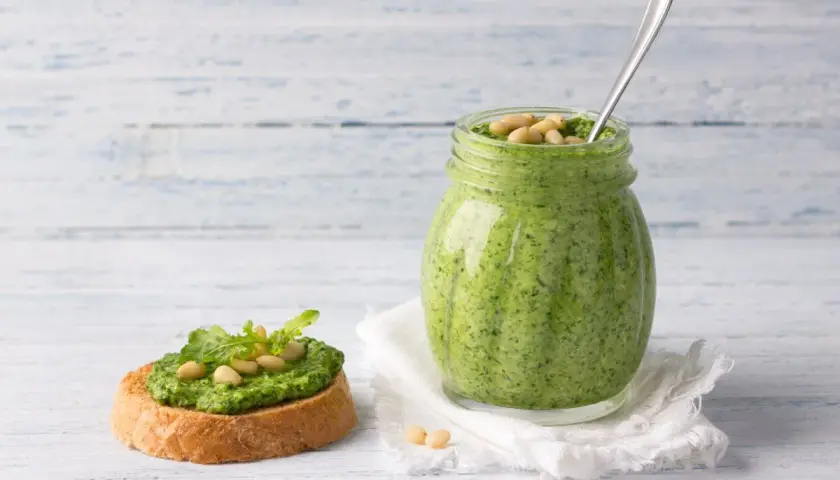 Pesto di rucola ricetta: un vasetto pieno di verde brillante e una bruschetta guarnita, pronti per essere gustati.