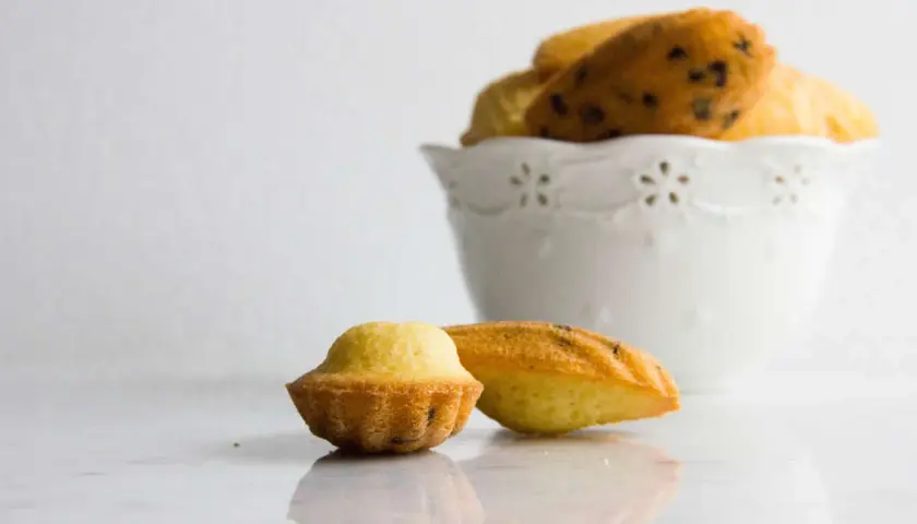 Due madeleine dolci, una a forma di conchiglia e una più tondeggiante, su un piano di marmo. Sullo sfondo, una ciotola bianca piena di altre madeleine.