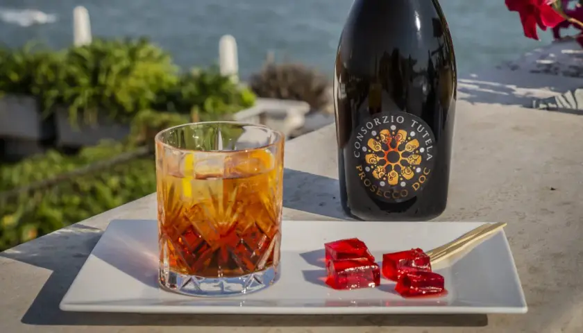Un bicchiere di spritz con spumante, una bottiglia di Prosecco e cubetti di gelatina rossa su un vassoio bianco, con vista sul mare.
