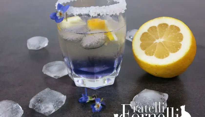 Rinfrescante liquore alla violetta spritz, guarnito con limone e fiori, circondato da cubetti di ghiaccio.