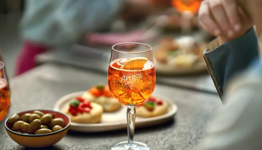 Un bicchiere di Aperol Spritz, ghiaccio e una fetta d'arancia, con olive e stuzzichini sullo sfondo.