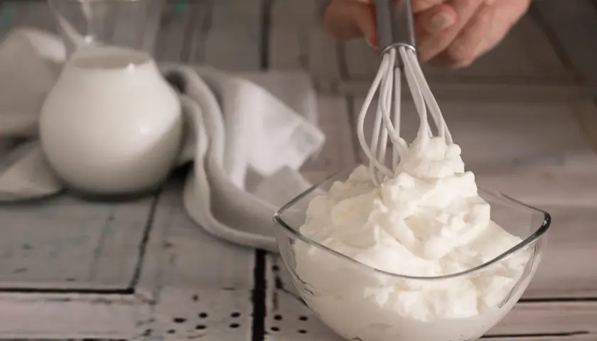 Una mano monta la panna in una ciotola di vetro. Sullo sfondo, una brocca di latte e un tovagliolo bianco.