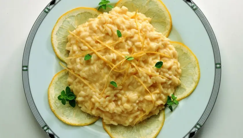 Un cremoso risotto al limone, guarnito con scorzette e foglioline di menta, circondato da fette di limone fresco.