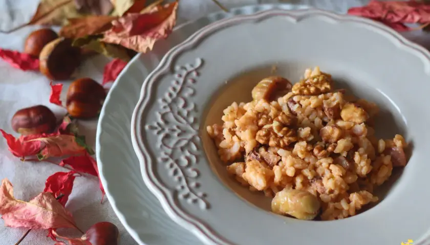 Un cremoso risotto castagne e gorgonzola, guarnito con noci e castagne intere, su un piatto decorato, circondato da foglie autunnali e castagne.