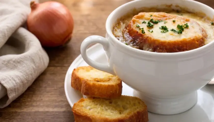 Una tazza di zuppa di cipolle fumante, con crostini dorati e formaggio fuso, accanto a una cipolla cruda e un tovagliolo di lino.