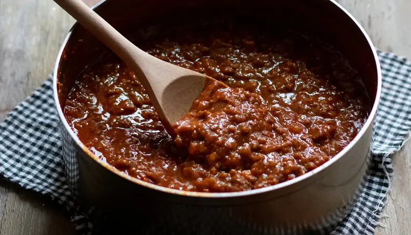 Un pentolino pieno di ragù alla bolognese, pronto per condire la pasta. Un cucchiaio di legno affonda nella salsa densa e profumata.
