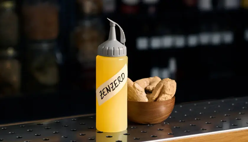 Flacone giallo con etichetta "ZENZERO" e una ciotola di radici di zenzero essiccate.