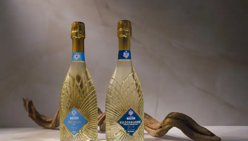 Due bottiglie di Prosecco Maschio, una DOC Treviso e l'altra Valdobbiadene DOCG, con un ramo secco. Scopri come si fa il Prosecco.