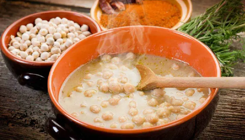 Una fumante zuppa di ceci in una ciotola arancione, con ceci secchi, spezie e rosmarino sullo sfondo.