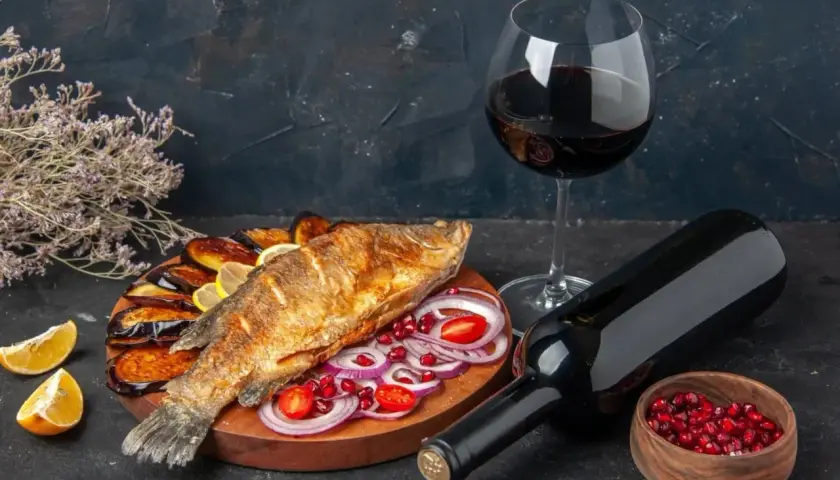 Un delizioso pesce fritto su un piatto con melanzane, cipolle rosse e pomodorini, accompagnato da vino rosso.