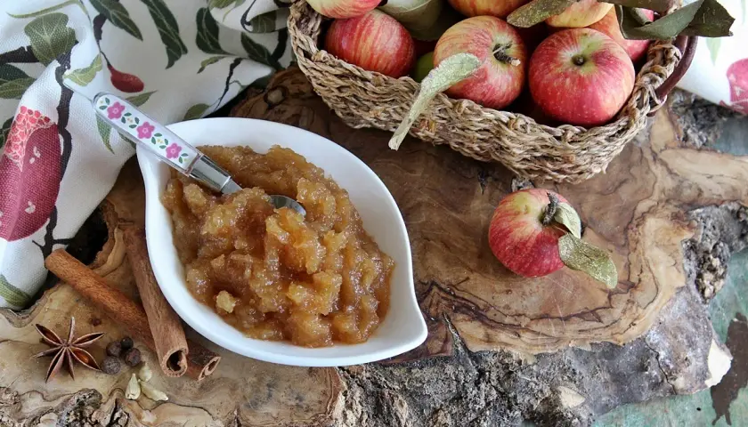 Una ciotola di composta di mele, profumata con cannella e anice stellato, perfetta per la marmellata di natale. Mele rosse in un cesto sullo sfondo.