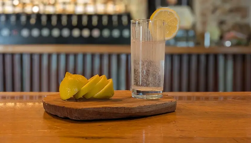 Un bicchiere di acqua frizzante con una fetta di limone, pronto per diventare la base perfetta per un rinfrescante spritz. Accanto, spicchi di limone su un tagliere di legno.
