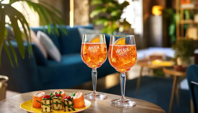 Due bicchieri di cocktail con Aperol Spritz, ghiaccio e fetta d'arancia, accompagnati da stuzzichini su un tavolino in legno.