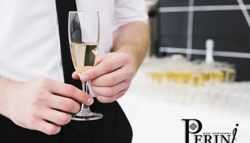 Un bicchiere di prosecco vino dorato, tenuto da mani eleganti, con una piramide di calici sullo sfondo.