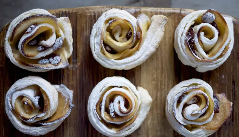 Sei rose di pasta sfoglia con mele e zucchero di canna, cosparse di zucchero a velo, su un tagliere di legno.