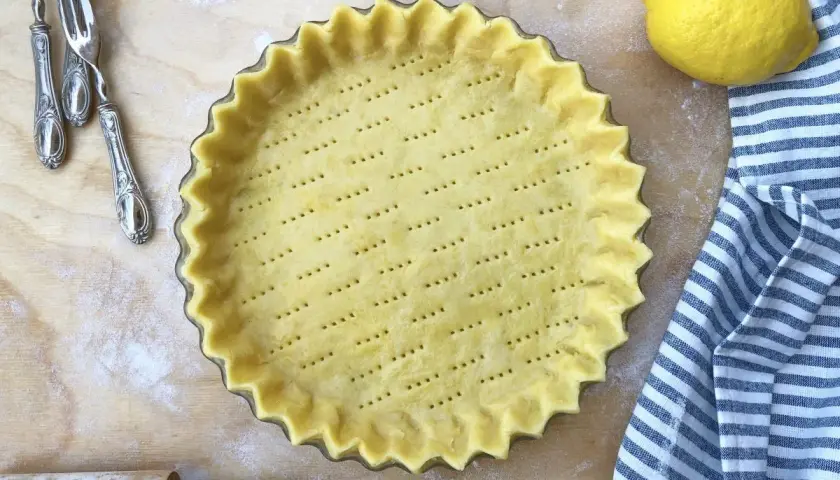 Base di pasta frolla pronta in teglia, forata e con bordi ondulati, accanto a un limone e un canovaccio a righe.