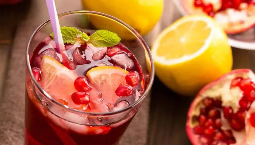 Un rinfrescante spritz al melograno con fette di limone, chicchi rossi e foglie di menta, accompagnato da limoni e melograni freschi.