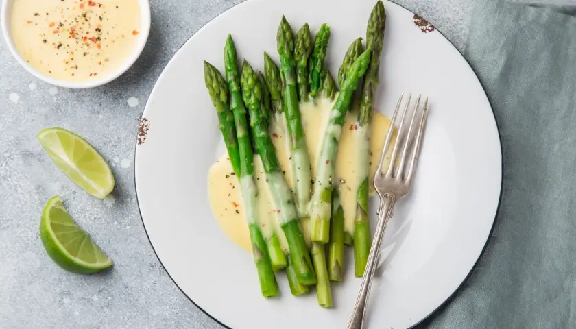Asparagi freschi conditi con una cremosa salsa olandese, accompagnati da spicchi di lime e una forchetta d'argento.