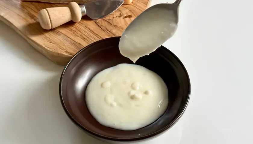 Un cucchiaio versa una densa crema di parmigiano in una ciotola. Sul tagliere, un pezzo di formaggio e un coltellino.