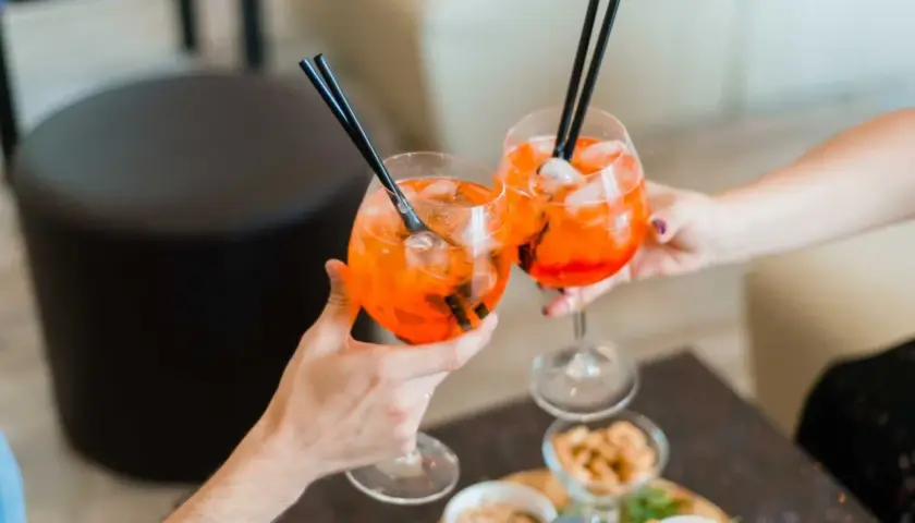 Due mani brindano con due spritz pieni di ghiaccio e cannucce nere. Sul tavolo, stuzzichini invitanti.