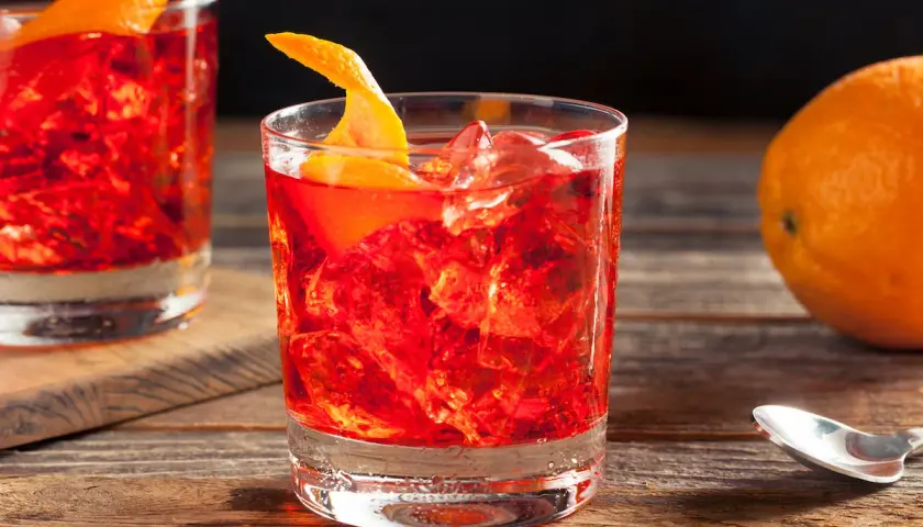 Un bicchiere di rosso vibrante, ghiaccio e scorza d'arancia. La ricetta americano è pronta per essere gustata, con un'arancia sullo sfondo.