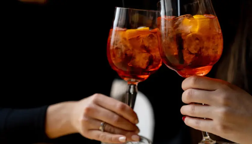 Due mani brindano con due spritz, un superalcolico rinfrescante con ghiaccio e arancia, su sfondo scuro.