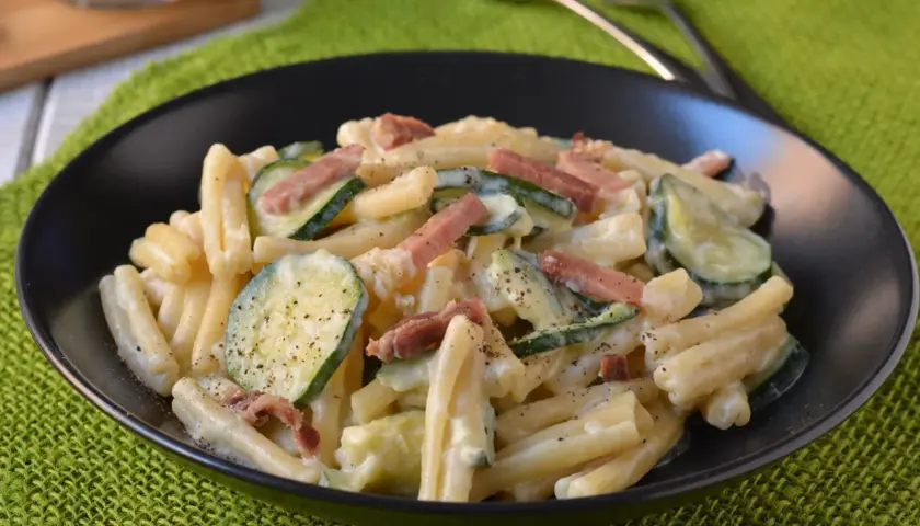 Piatto di pasta cremosa con zucchine a rondelle e cubetti di speck croccante, guarnito con pepe nero.