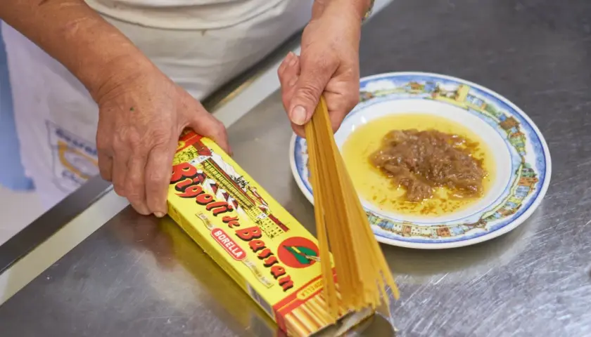Mani che preparano i bigoli in salsa, con la scatola di pasta e un piatto di salsa di acciughe e cipolle.