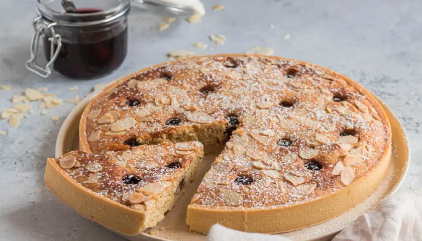 Torta con crema frangipane, mandorle a lamelle e ciliegie, spolverata di zucchero a velo. Accanto, un vasetto di confettura.