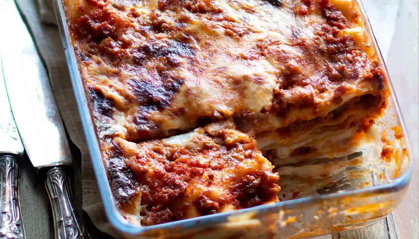 Lasagne alla bolognese appena sfornate, dorate e filanti, in una teglia di vetro. Accanto, due coltelli d'argento.