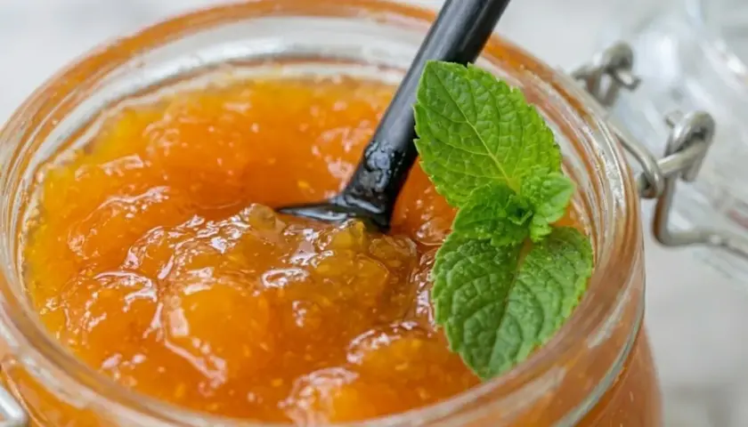 Vasetto di vetro pieno di marmellata di albicocche fatta in casa, con un cucchiaio nero e una fogliolina di menta fresca.
