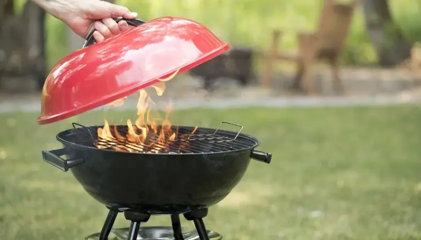 Mano solleva il coperchio rosso di un barbecue a carbonella con fiamme vive. Perfetto per chi vuole sapere se posso usare legna nel barbecue a carbonella.
